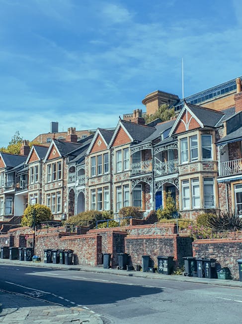 A row of Victorian-style terraced houses situated on a city street under a clear blue sky. The residences feature ornate white decorative ironwork railings and trim, with large bay windows on the uppe