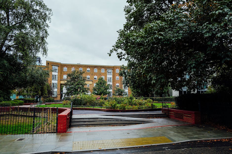 The image depicts a residential street scene during daytime, showing the exterior of a multi-storey brick apartment building in the background with numerous white-framed windows and balconies. In the foreground, there is a wooden ramp with metal handrails on each side, leading down from a paved sidewalk to a lower level of paved outdoor space. The pavement is wet, suggesting recent rain, and is surrounded by greenery, including large trees with dense foliage on both sides, and a landscaped garden area with flowering plants and bushes. The scene appears to be set in an urban environment, suitable for house and apartment removals, with movers VBS Belsize Park potentially utilizing this area for loading or unloading furniture and boxes during a home relocation process. The lighting is natural and diffused under an overcast sky, capturing the typical atmosphere of a city neighbourhood.