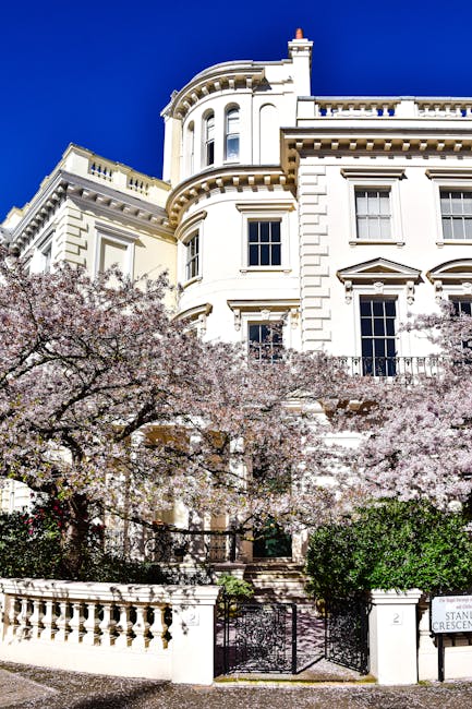 A tall, white, multi-storey residential building with ornate architectural details, including curved bay windows and decorative cornices, set against a clear blue sky. In front of the building, there are blooming pink cherry blossom trees and green shrubs, with a black metal gate and white concrete balustrade at the entrance. The scene suggests an upscale residential area in Belsize Park, suitable for home relocation or moving services, with the building possibly being a target for furniture transport or interior packing and moving activities, as orchestrated by Movers Belsize Park. The overall lighting is bright, emphasizing the building's exterior features and the vibrant flowering trees.