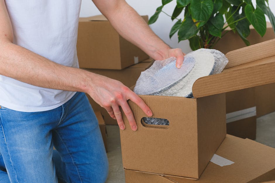 Two movers from Movers Belsize Park are inside a well-lit room with white walls and hardwood flooring, preparing for a home relocation. The worker on the left, with curly black hair and a beard, is carrying two stacked medium-sized cardboard boxes, with red packing tape securing them and black printed labels indicating their size. The worker on the right, wearing a headband, is holding a single cardboard box, engaging in a conversation about the packing and moving process. Behind them, three additional cardboard boxes are positioned on the floor near a doorway, ready to be loaded into a moving van. The room features two large, arched windows allowing natural light to illuminate the space, and a vintage green upholstered armchair is visible to the right, alongside the doorway. The scene captures the loading process of furniture transport typical of house removals, with packing materials and equipment such as trolleys or straps not visible, emphasizing the careful handling of boxes by professional movers during a house move, as detailed on the Belsize Village removals tips page.
